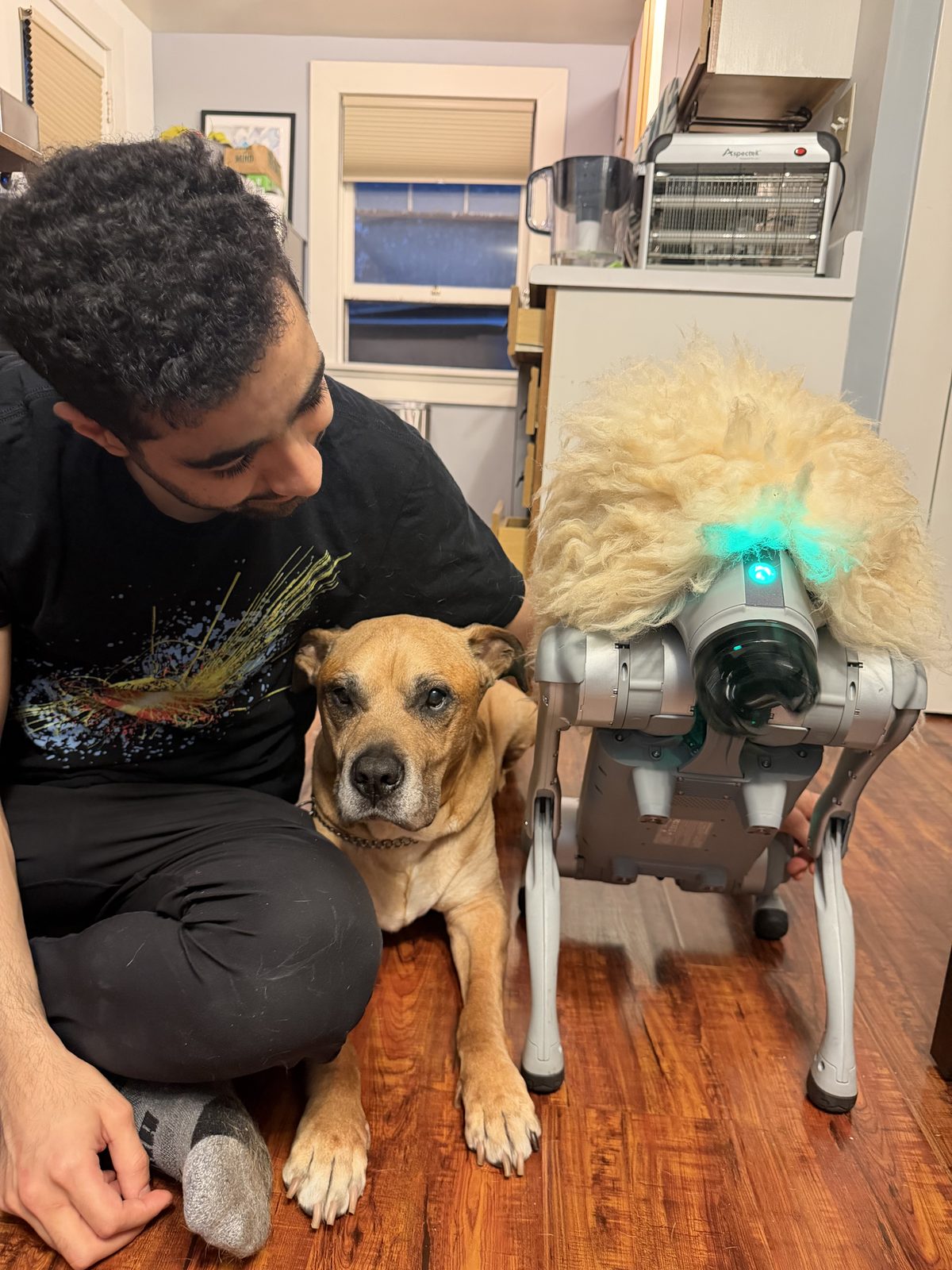 Ephraiem seated on the floor with a dog and Claudius Maximus, the quadruped robot in a fluffy mane
